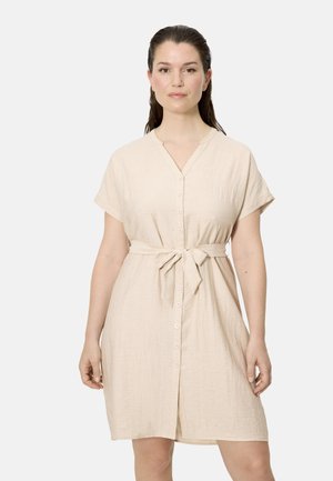 Mujer de cabello oscuro recogido, vestida con un vestido beige de manga corta con botones y cinturón en la cintura, de pie frente a un fondo claro y sencillo.