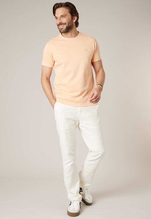 Lachende man met baard en middellang haar draagt een perzikkleurig shirt met korte mouwen, witte broek en witte sneakers, staand met één hand in de zak.