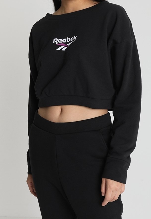 Sudadera corta negra con cuello redondo y mangas largas, que presenta un logo de Reebok en púrpura y blanco en el frente. Textura de tela suave.