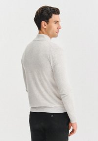 Jasnoszary sweter z kołnierzykiem, prążkowanymi mankietami i dołem, wykonany z miękkiego, teksturowanego materiału. Noszony z ciemnymi spodniami, widok z tyłu.