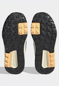 Zapatillas de trail running con suelas de goma negra texturizada, que presentan acentos en marrón claro, múltiples patrones de tracción y una entresuela beige suave.