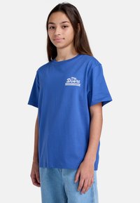 Camiseta de algodón azul con mangas cortas y escote redondo. Presenta un logo impreso en blanco en la parte izquierda del pecho. Combinada con unos pantalones vaqueros de mezclilla clara.