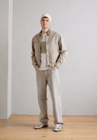 Chaqueta beige con cuello doblado, camiseta de cuello redondo gris claro y jeans de mezclilla claros, combinados con zapatillas verdes y blancas. Los accesorios incluyen un gorro blanco.