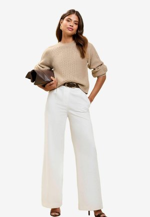 Beige cable knit sweater, white wide-leg trousers, brown belt, and clutch bag. High-heeled sandals complete the outfit. Smooth textures throughout.