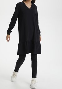 Robe noire à manches longues avec col en V et ourlet volanté, associée à des leggings noirs et des baskets blanches, présentée en position debout.
