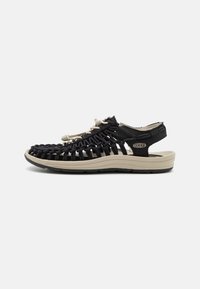 Black woven sandal with a beige rubber sole, featuring adjustable straps and a textured upper for breathability and comfort.