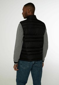 Czarna kamizelka puchowa z wysokim kołnierzem, pikowanym wzorem i dopasowanym krojem, noszona na szarej koszulce z długim rękawem i niebieskich jeansach.