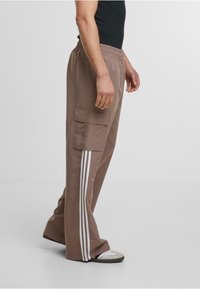 Bruna cargobyxor med elastisk midja och två sidofickor, med tre vertikala vita ränder längs yttre sömmarna. Slät tygtextur.