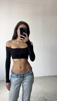 Černý top s odhalenými rameny a dlouhými rukávy, s uzlem na přední straně, v kombinaci se světle modrými džíny. Hladká tkanina a uvolněný střih.