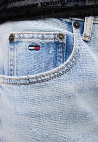 Jeans de mezclilla azul claro con un bolsillo superior visible, que cuentan con un pequeño parche de logo rojo, blanco y azul, y detalles de hardware en cobre.