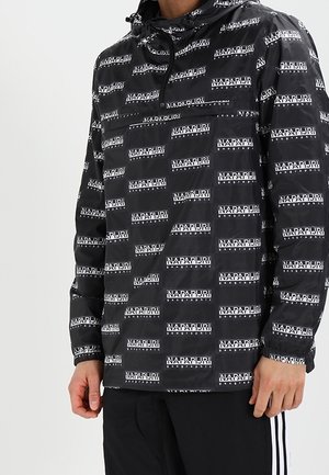 Sort hættetrøje med gentagne hvide Napapijri-logoer båret af en person med sorte bukser med hvide sidelænker.