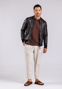 Zwarte leren bomberjack over een bruin gestructureerd polo, gecombineerd met lichtbeige broeken en bruine sandalen. Eenvoudige, urban outfit.