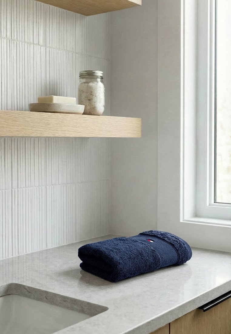 Serviette bleu marine pliée sur un comptoir en marbre à côté de l'évier, étagère flottante en bois avec porte-savon et bocal, mur en carrelage blanc texturé, fenêtre à proximité.