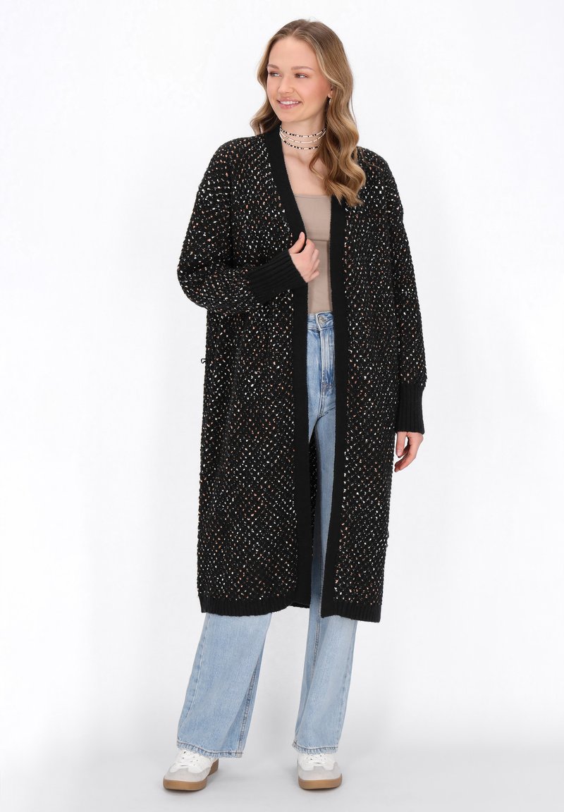 Lång svart cardigan med en lös passform, texturerad stickning som har multikolorade fläckar, ribbade ärmslut och rakt nederkant. Bärs över en beige topp och jeans.