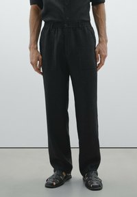 Pantalon noir, coupe ample avec une taille élastique, doté de deux poches avant et d'un tissu doux et texturé. Associé à des sandales tressées noires.
