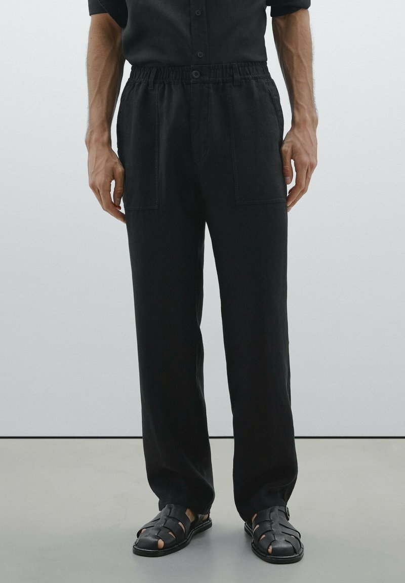 Pantalon noir, coupe ample avec une taille élastique, doté de deux poches avant et d'un tissu doux et texturé. Associé à des sandales tressées noires.