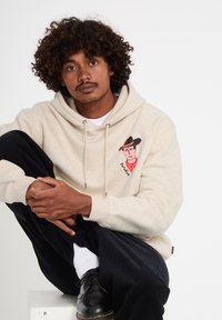 Homme aux cheveux bouclés portant un sweat à capuche beige avec un motif de cow-boy, pantalon noir, chaussettes blanches et chaussures noires, assis en tailleur.