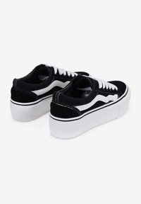 Zapatillas de ante negro con cordones blancos, que presentan un diseño de ondas blancas distintivo y una suela gruesa blanca. La vista trasera muestra los detalles de costura.