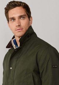 Chaqueta verde oscuro con cuello alto, cierres de botón a presión y bolsillo lateral. Presenta un cuello marrón en contraste y una etiqueta con la marca en la manga.