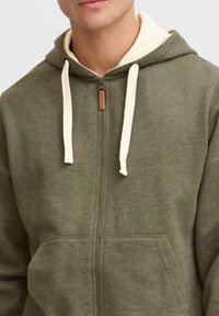 Olive green zip-up hoodie with a cream-colored inner lining, adjustable drawstrings, and front pockets. Textured fabric with visible seams.