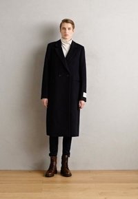 Manteau en laine noir à double boutonnage avec un col cranté, poches latérales et un accent blanc sur la manche, associé à des bottes à lacets marron.