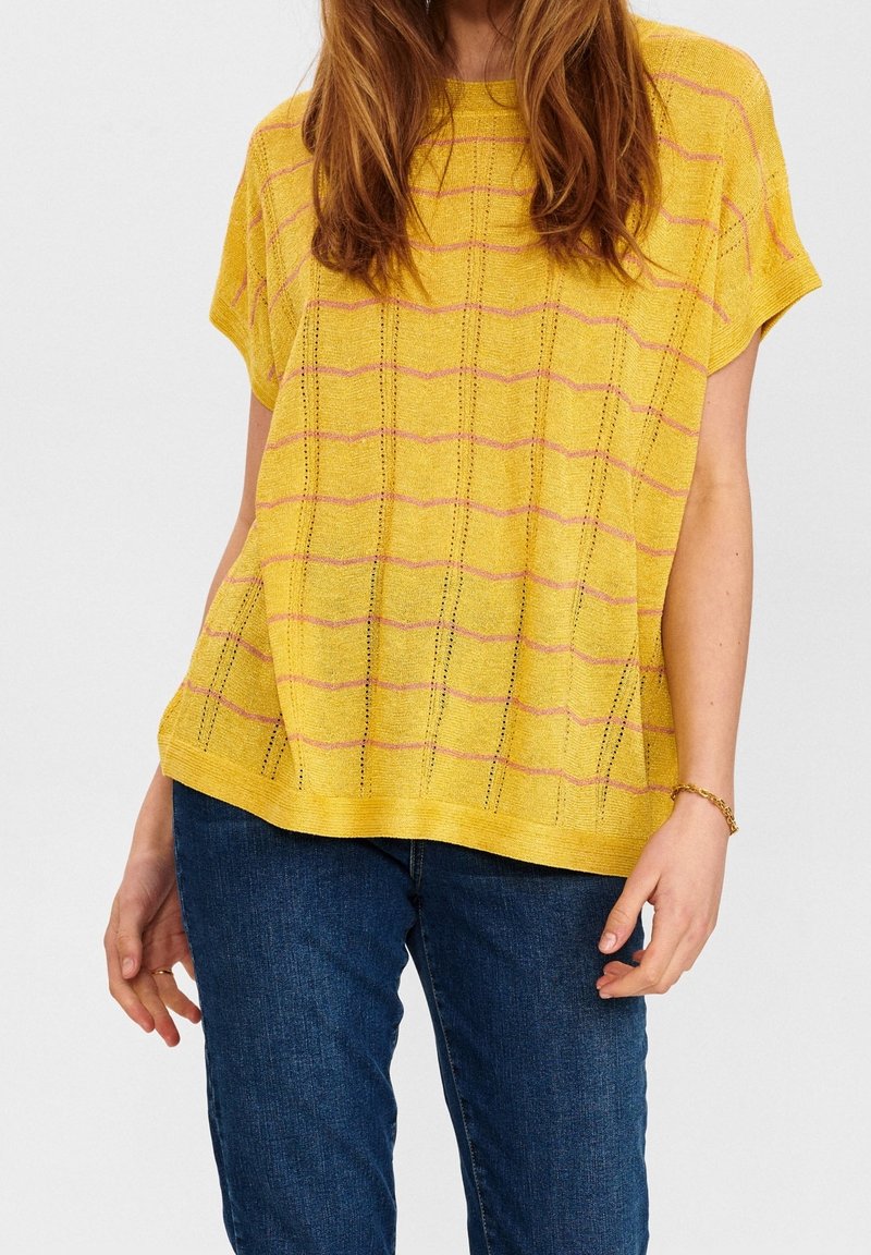 Gul stickad topp med korta ärmar, med ett strukturerat mönster av rosa ränder och små öppna stygn, kombinerad med mörkblå jeans.