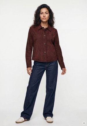 Frau steht und trägt eine braune, geknöpfte Jacke, dunkelblaue Jeans mit weitem Bein und weiße Sneakers vor einem schlichten weißen Hintergrund.