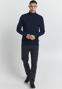 Mornarsko pulover s turtleneck ovratnikom in rebrasto teksturo, v kombinaciji s temnimi hlačami in črnimi športnimi copati, predstavlja oprijet silhueto.