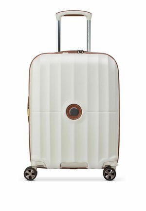Valise rigide blanche avec texture nervurée, accents marron, poignées rétractables et quatre roues. Présente un logo rond à l'avant.