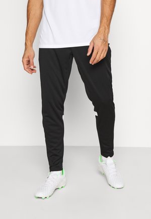 Zwarte sportbroek met een taps toelopende pasvorm, voorzien van witte accenten aan de zijkant. Draagt witte en groene voetbalschoenen. Gladde stoftextuur.