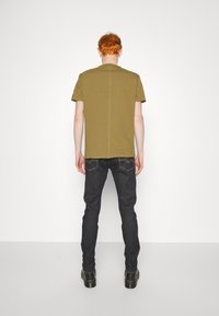 Camiseta de algodón verde oliva de manga corta, con una costura central en la parte posterior, combinada con jeans oscuros y botas negras.