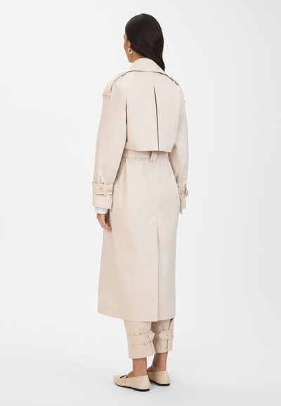 Trench coat beige con ampio colletto, vita con cintura e polsini abbottonati. Tessuto liscio con vestibilità sartoriale, dotato di uno spacco sul retro per il movimento.