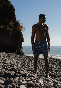 Zwemshorts in donkerblauw met een palmbladpatroon, gemaakt van lichtgewicht stof. Model staat op een kiezelstrand bij het water.