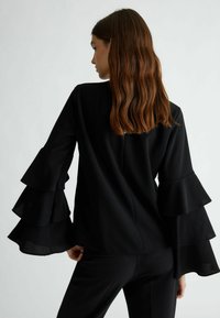 Top nero con maniche a sbuffo e a strati. Tessuto morbido, vestibilità tailored e dettaglio della cucitura posteriore. Indossato con pantaloni neri coordinati.