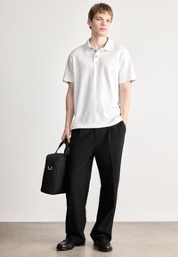 Bílé polo tričko s krátkým rukávem, černé kalhoty a černé boty. Drží černou tašku s držadlem, na jednobarevném pozadí.