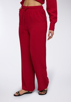 Pantaloni a gamba larga rossi in tessuto sgualcito, con cintura elastica dotata di cordino e tasche laterali. Abbinati a sandalini neri.