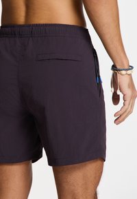 Férfi sötétlila fürdőnadrág, amely könnyű anyagból készült, elasztikus derékrésszel, hátsó zsebbel és cipzáras oldalzsebbel rendelkezik.