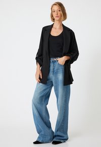 Blazer negro con mangas arremangadas sobre una camiseta de tirantes negra, combinado con jeans de mezclilla azul de pierna ancha y cintura alta. Zapatos negros completan el atuendo.