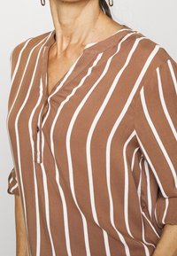 Chemise marron à manches longues avec des rayures blanches verticales, patte de boutonnage et col en V. Tissu doux avec une coupe décontractée.