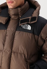 Brun och svart isolerad pufferjacka med hög krage, kardborreaccenter och en vit logotyp på bröstet. Har en quiltad textur.