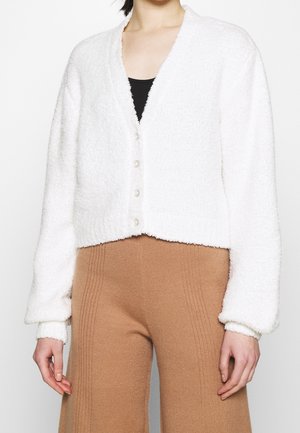 Femme portant un cardigan blanc duveteux boutonné sur un top noir et un pantalon côtelé camel taille haute.