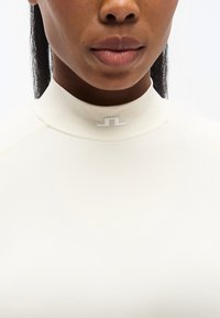 Εκρού ρούχο με ψηλό γιακά και απαλό ύφασμα, με μικρό λευκό λογότυπο στον γιακά. Διαθέτει ενισχυμένες ραφές για ανθεκτικότητα.