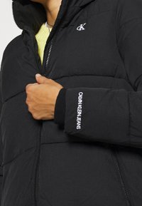 Czarna kurtka puchowa z zamkiem z przodu, z kapturem i logo na klatce piersiowej oraz rękawie. Materiał wydaje się pikowany, co zapewnia izolację.