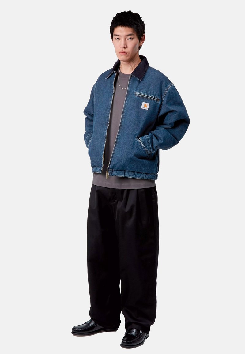 Giubbotto di jeans con una finitura blu, colletto a contrasto, tasche frontali e un'etichetta con marchio. Abbinato a una camicia grigia e pantaloni plissettati neri.