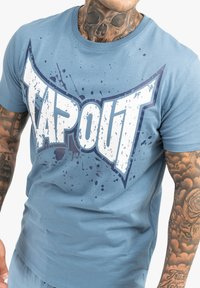 Mėlynas medvilnės marškinėlis su dideliu baltu "TAPOUT" grafiku, kuris atrodo nusidėvėjęs, akcentuotas violetiniais purslų raštais. Trumpomis rankovėmis ir apvaliu kaklu.