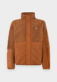Brown jacket made of plush fleece and quilted fabric. Features a high collar and a small logo on the upper chest. Textured accents throughout.