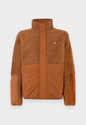 Brun jakke laget av myk fleece og quiltet stoff. Har høy krage og en liten logo på øvre bryst. Teksturerte detaljer gjennom hele jakken.