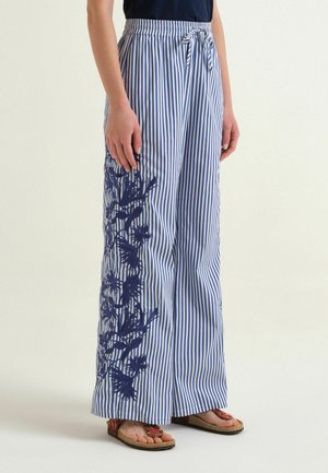 Donna che indossa pantaloni a gamba larga a righe verticali blu e bianche con stampa floreale blu scuro sul lato e vita annodata, abbinati a sandali.