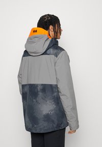 Veste imperméable grise et bleu marine avec capuche, présentant une accent orange sur le col et un design texturé.