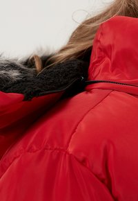Rød isoleret jakke med en krave i sort faux fur, glat overflade og lynlåslukning med teksturerede stofdetaljer.